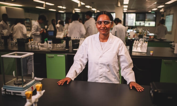 Woman in lab coat standing in front of a laboratory workspace with students working in the background.